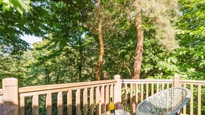 A wooden deck with wicker chairs and a small round table, surrounded by dense trees. - Fern View - Kelling Heath Holiday Park (Holt)