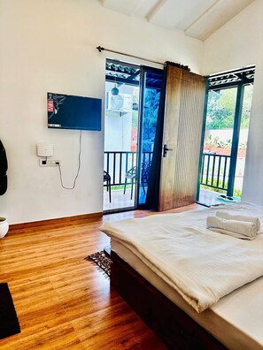 A bedroom with a bed, a TV mounted on the wall, and a sliding glass door leading to an outdoor area. - Casa Mounterria (Alibag)
