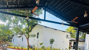 Uma área externa com mesas e cadeiras, um telhado coberto e um edifício ao fundo.