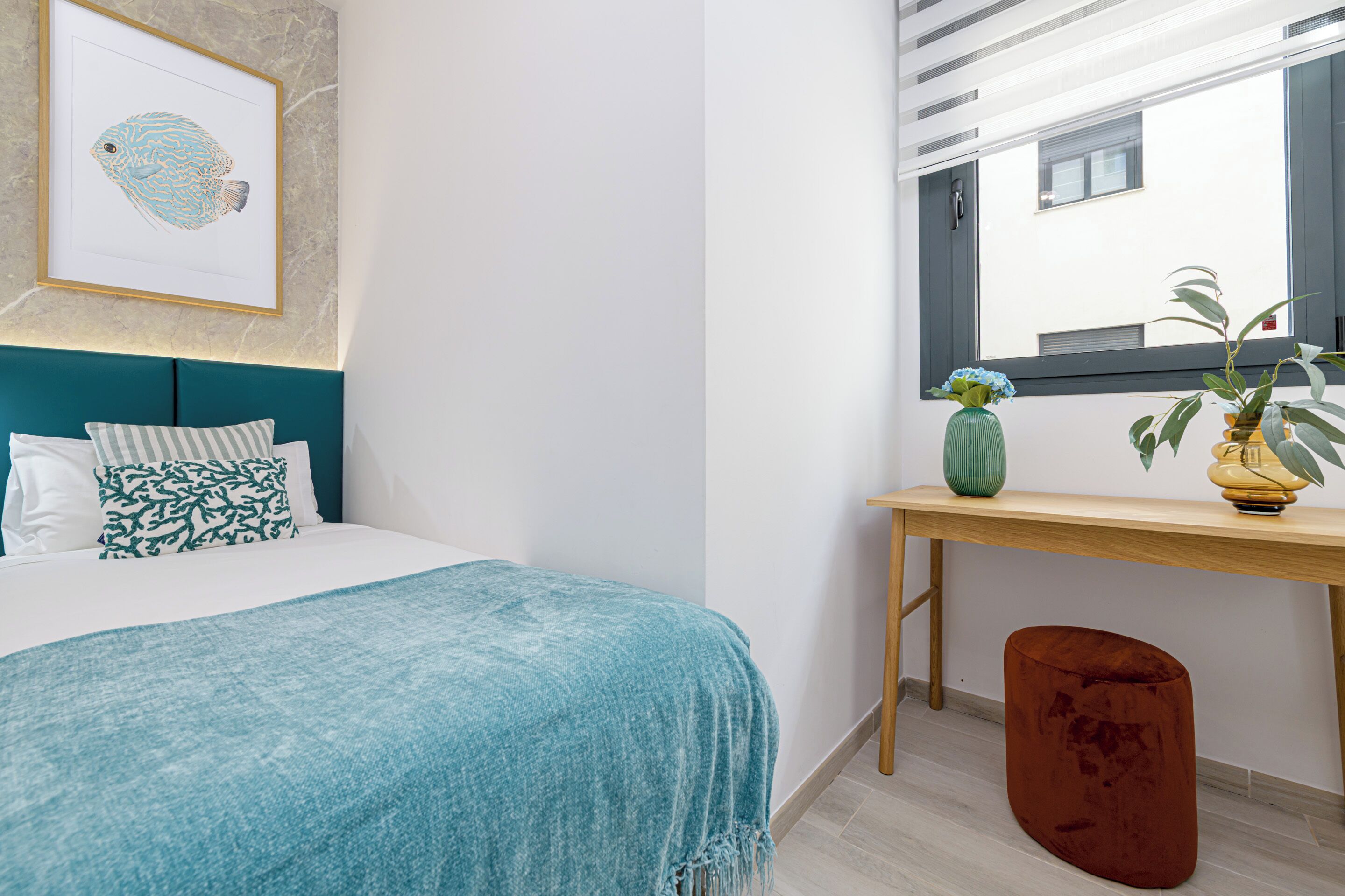 Un dormitorio con cama, una mesita de noche con un florero y planta, una ventana con persianas y un cuadro en la pared.