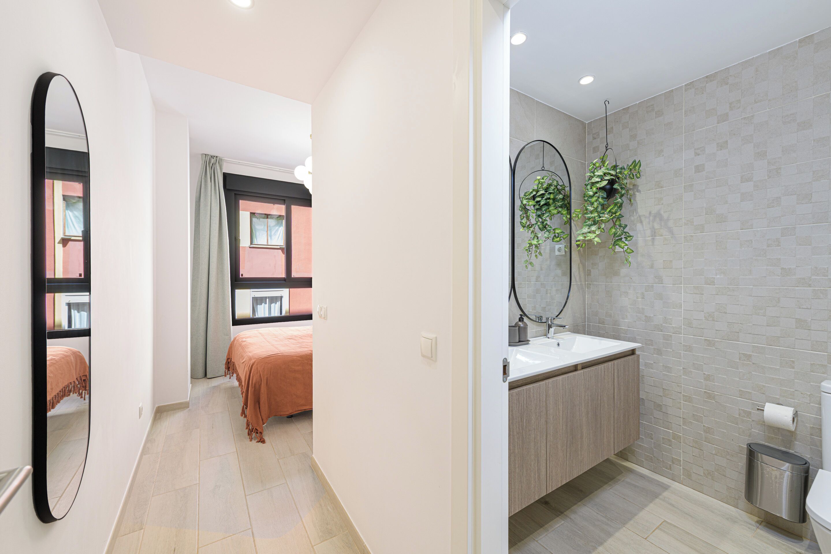 Une chambre d’hôtel moderne, dotée d’un grand miroir, d’un lit, d’une salle de bain avec un lavabo et des toilettes, et d’un couloir menant à une autre chambre.