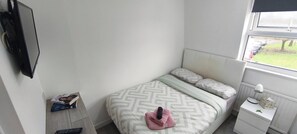 A bedroom with a bed, a bedside table, a nightstand, a TV mounted on the wall, and a window with a view of the outdoors. - Room-cozy Small Room in City Centre and (Chelmsford)