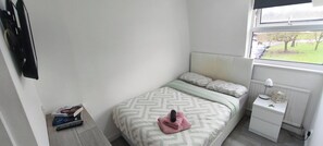 A bedroom with a bed, a nightstand, a lamp, a window with a view of trees, and a book on the nightstand. - Room-cozy Small Room in City Centre and (Chelmsford)