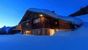 Uma cabana de madeira com telhado inclinado, rodeada por neve, e iluminada por luzes externas.