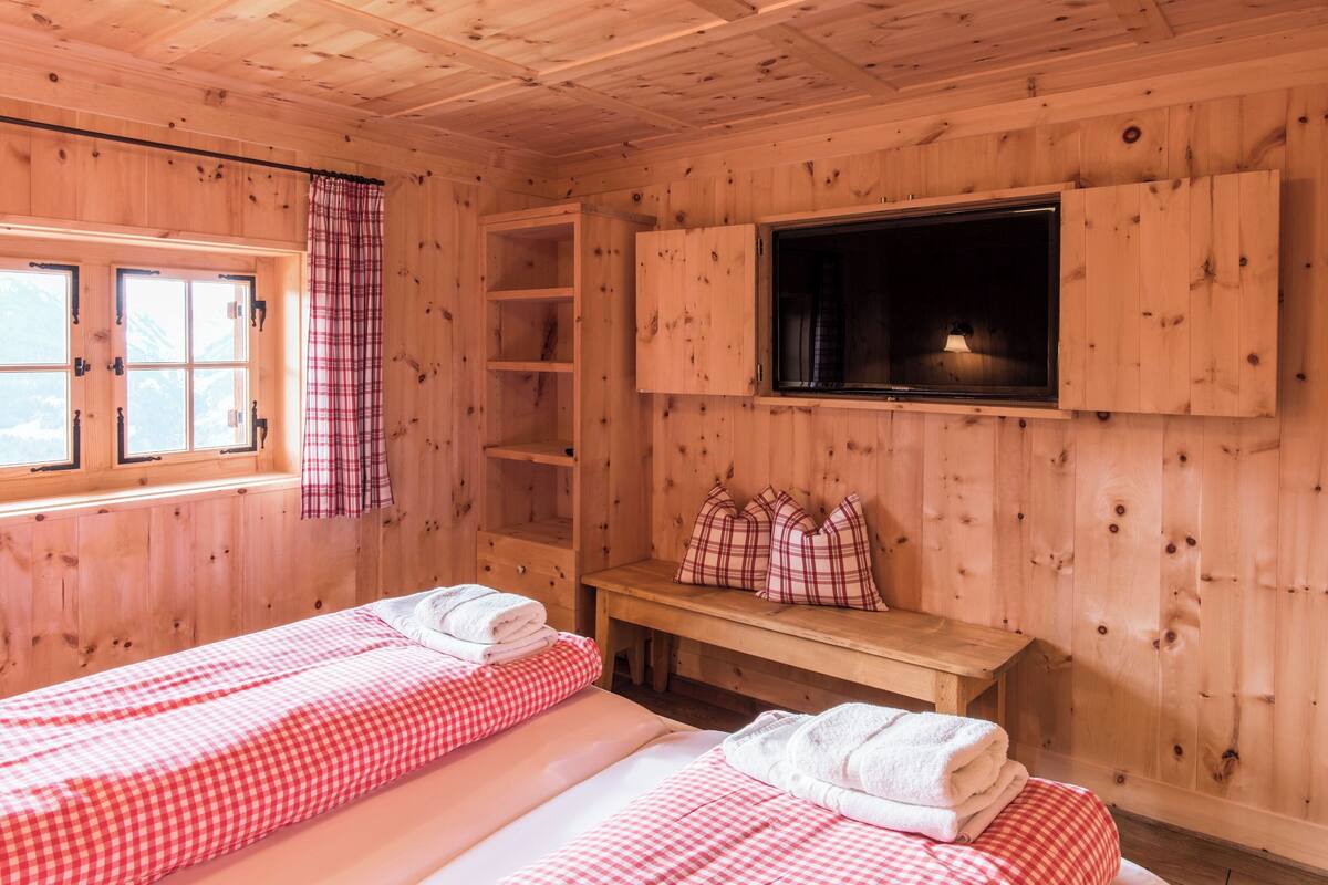 A wooden cabin room with two beds, a bench, a TV, and a window with a view.