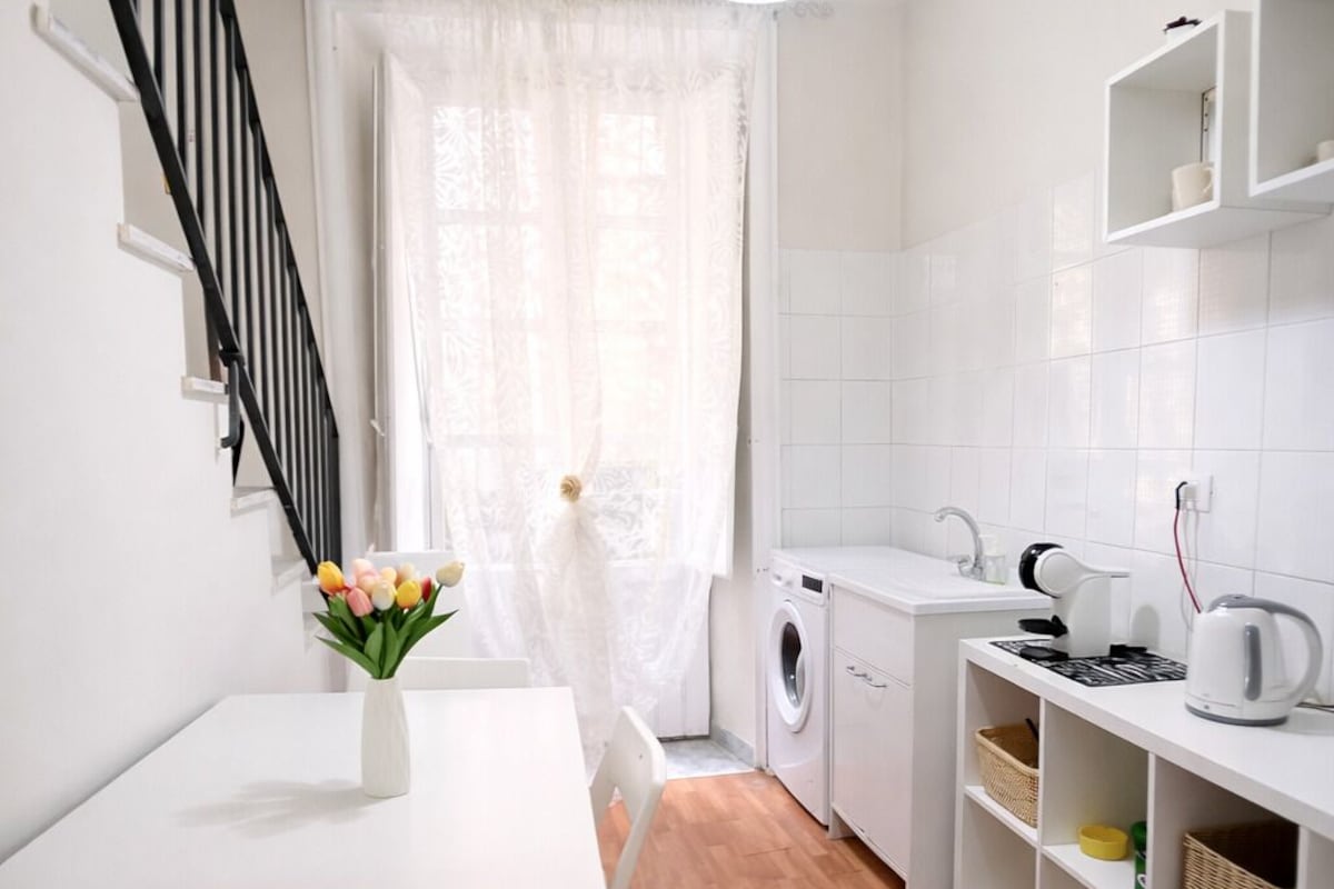 Una cucina compatta con lavatrice, un tavolo da pranzo con un vaso di fiori, una scala e scaffali bianchi.