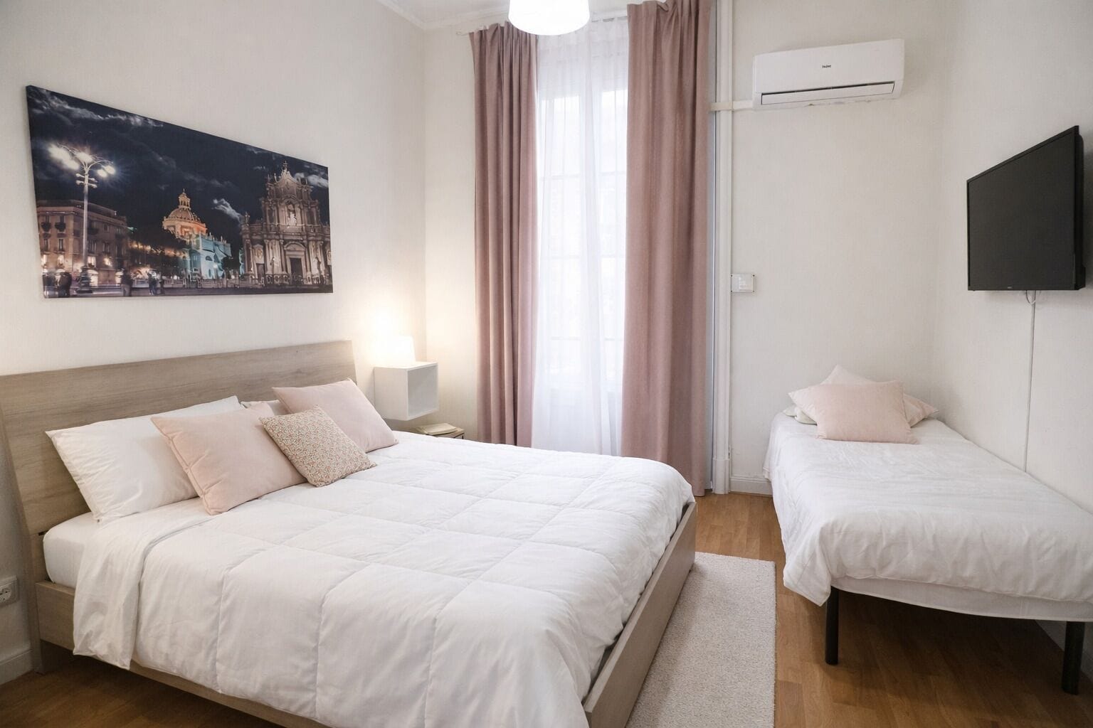 A hotel room with a large bed, a smaller bed, a flat-screen TV, an air conditioning unit, and a cityscape painting on the wall.
