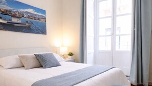 Un dormitorio con cama, mesita de noche, lámpara, ventana con cortinas y un cuadro en la pared.