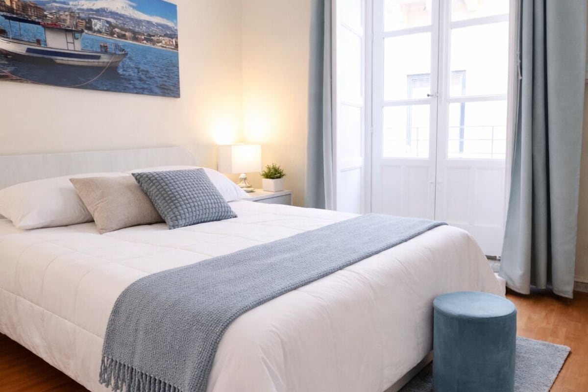 Una camera da letto con un letto, un comodino, una lampada, una finestra con tende e un quadro appeso al muro.