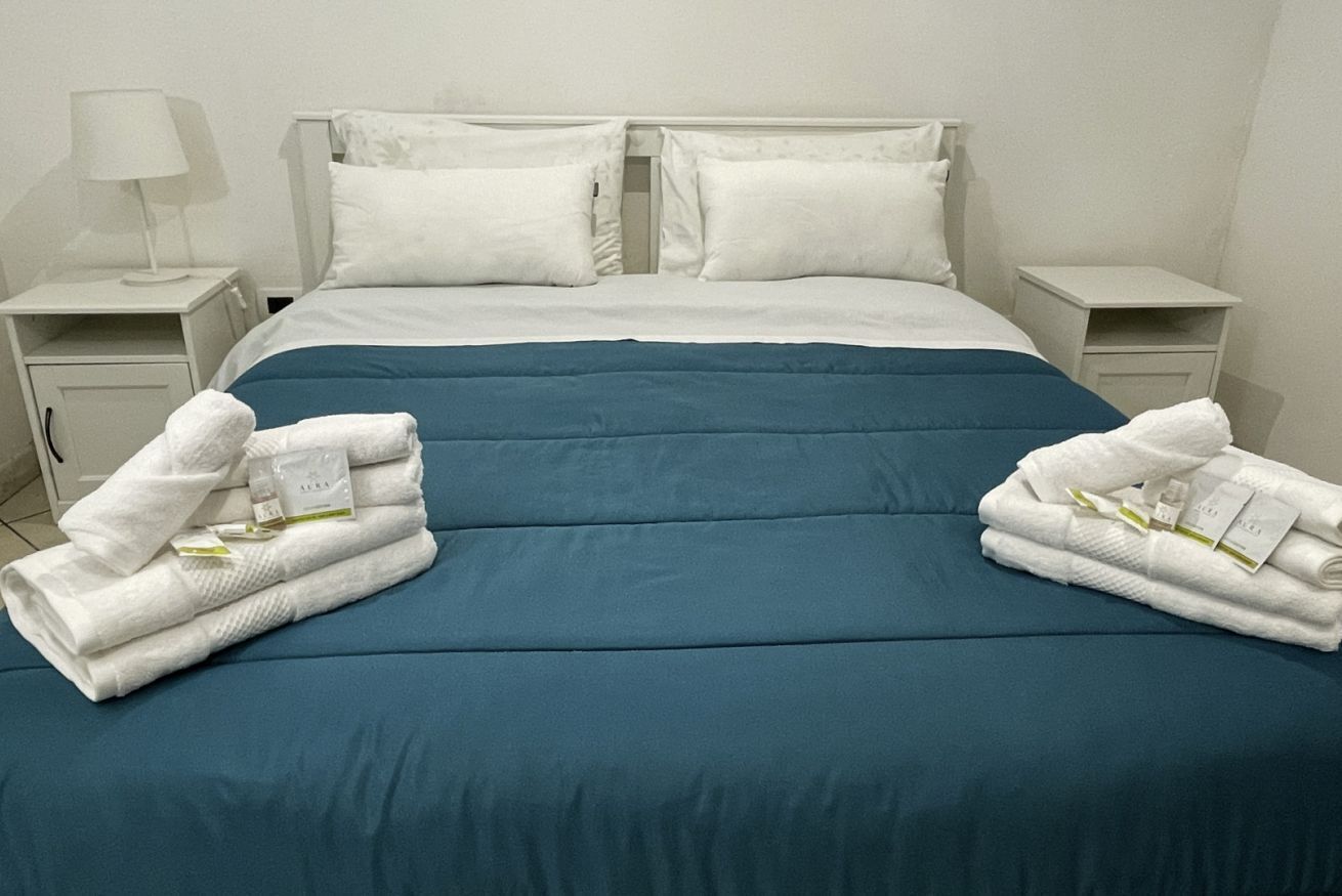Una cama bien hecha con una colcha azul, almohadas blancas y dos toallas blancas dobladas a cada lado.