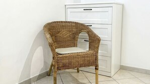 Una silla de mimbre con asiento y respaldo acolchados, ubicada en un rincón junto a un armario blanco y piso de baldosas.