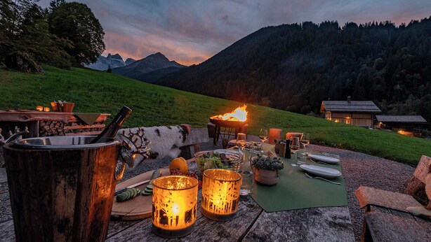 A rustic outdoor dining setup with a fire pit, wooden table, and mountain backdrop. - Berghaus Gauertal (Tschagguns)