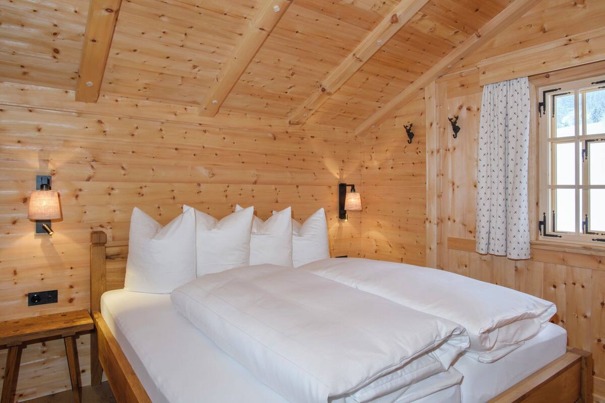 A wooden cabin room with a bed, wall-mounted lamps, and a window with curtains.