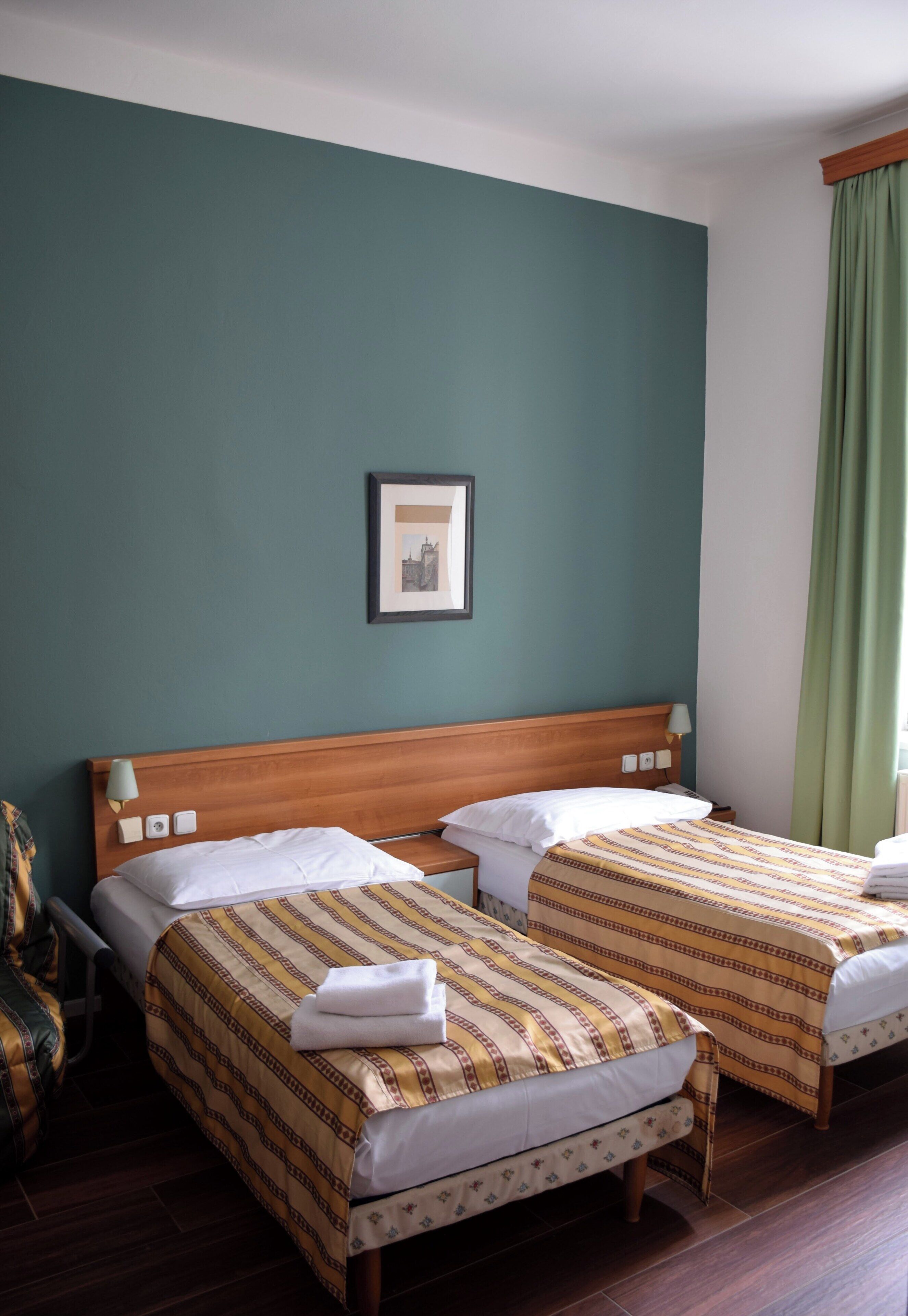 A hotel room with two single beds, a wooden headboard, a framed picture on the wall, and a green curtain.