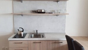 Una cocina moderna con estufa integrada, pileta y armarios de madera. Hay dos estantes sobre la encimera, uno con una olla y vasos, y el otro vacío. Una silla negra está ubicada a la derecha.