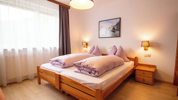 Een tweepersoonsbed met houten hoofdeinde en voeteinde, twee nachtkastjes met lampen, een raam met dunne gordijnen en een ingelijste foto aan de muur.