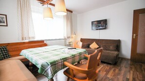 Een kamer met een bed, een houten stoel, een tafel met een groen geruit tafelkleed, een televisie aan de muur en een raam met gordijnen.