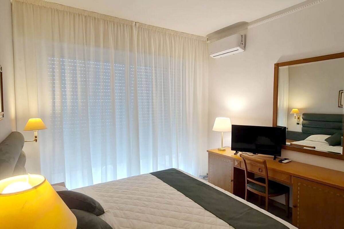 A hotel room with a bed, a desk with a television, a chair, a lamp, and a large window with curtains.