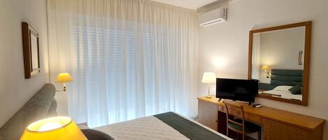 A hotel room with a bed, a desk with a television, a chair, a lamp, and a large window with curtains.
