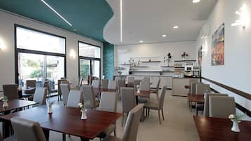 A modern dining area with tables and chairs, a kitchen in the background, and large windows.