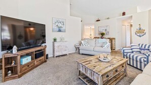 Een woonkamer met een grote flatscreen televisie, een houten tv-meubel, een lichtkleurige bank, een gestreepte bank en een salontafel.