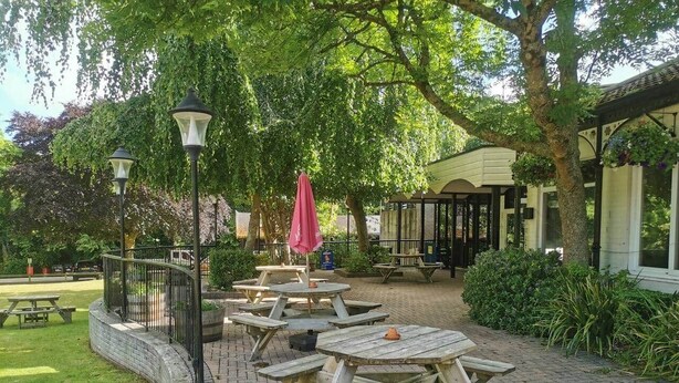 Ein gepflasterter Au ßenbereich mit Picknicktischen und einem roten Sonnenschirm, umgeben von Bäumen und einem Gebäude mit überdachter Terrasse.