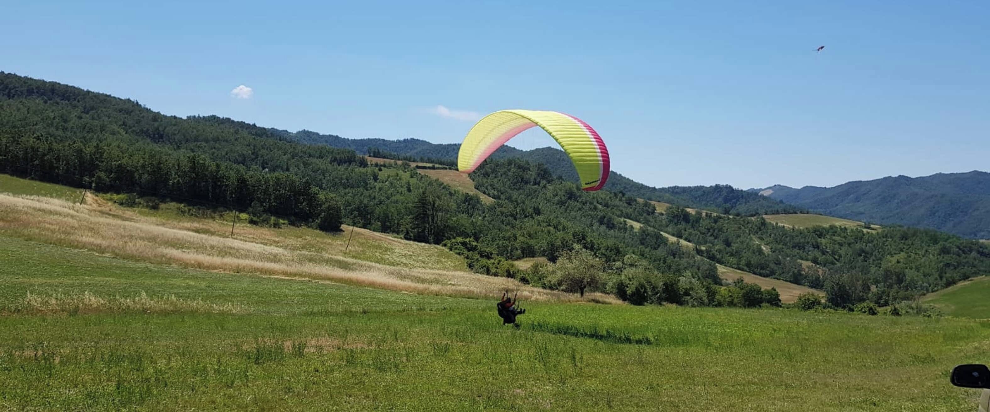Une personne pratiquant le parapente au-dessus d’un champ herbeux, avec des collines en arrière-plan.