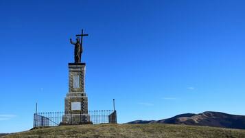 Una estatua sobre un pedestal con una cruz, rodeada por una valla, frente a un cielo azul despejado y montañas distantes.