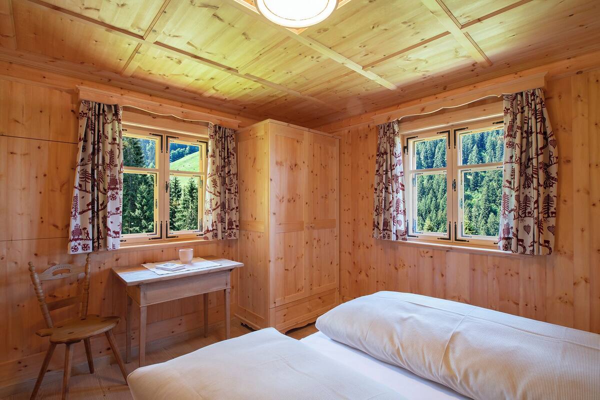 Una habitación de cabaña de madera con dos ventanas, un escritorio, una silla y una cama.