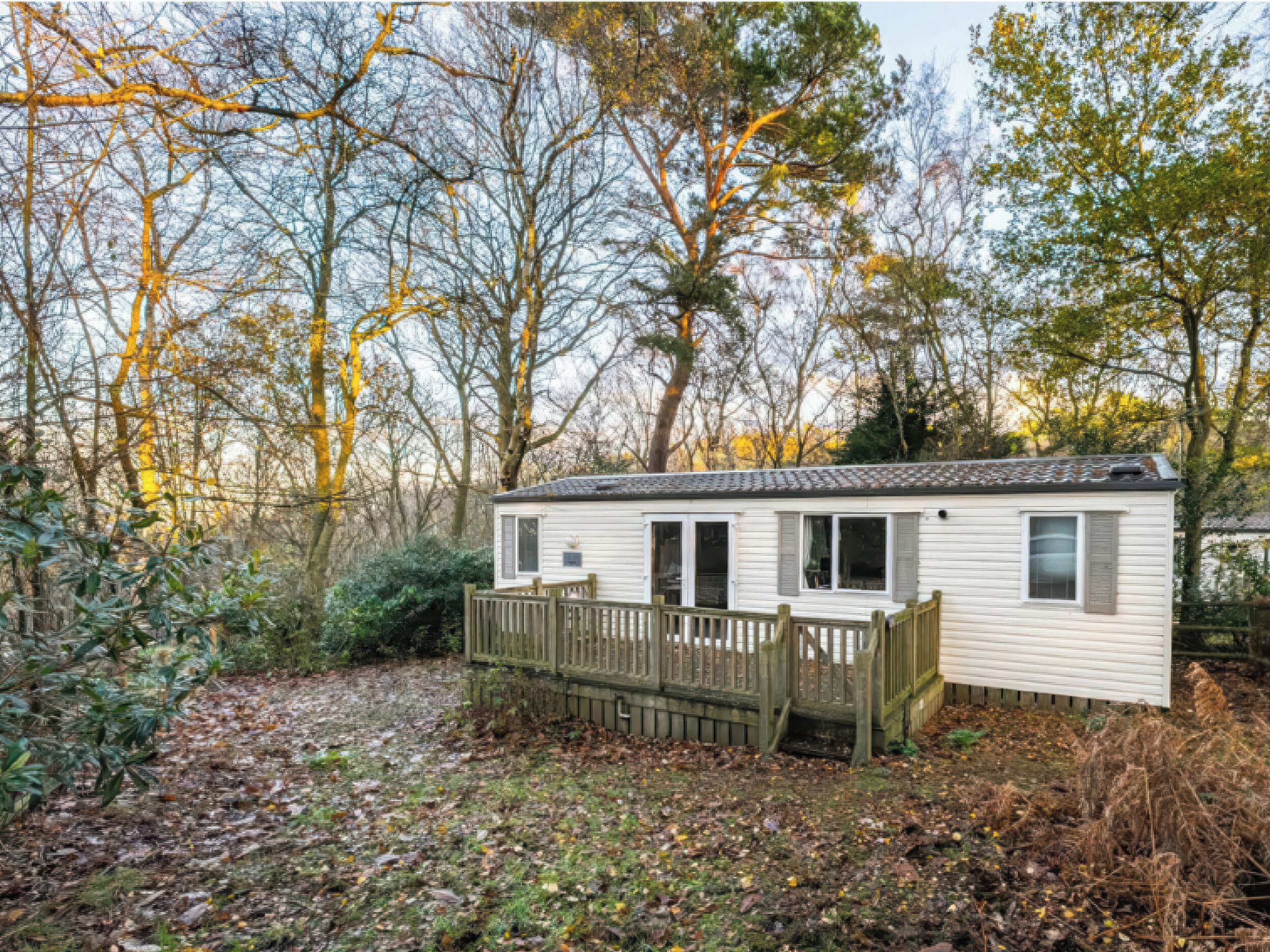 A mobile home with a deck and a fenced yard, surrounded by trees and vegetation.