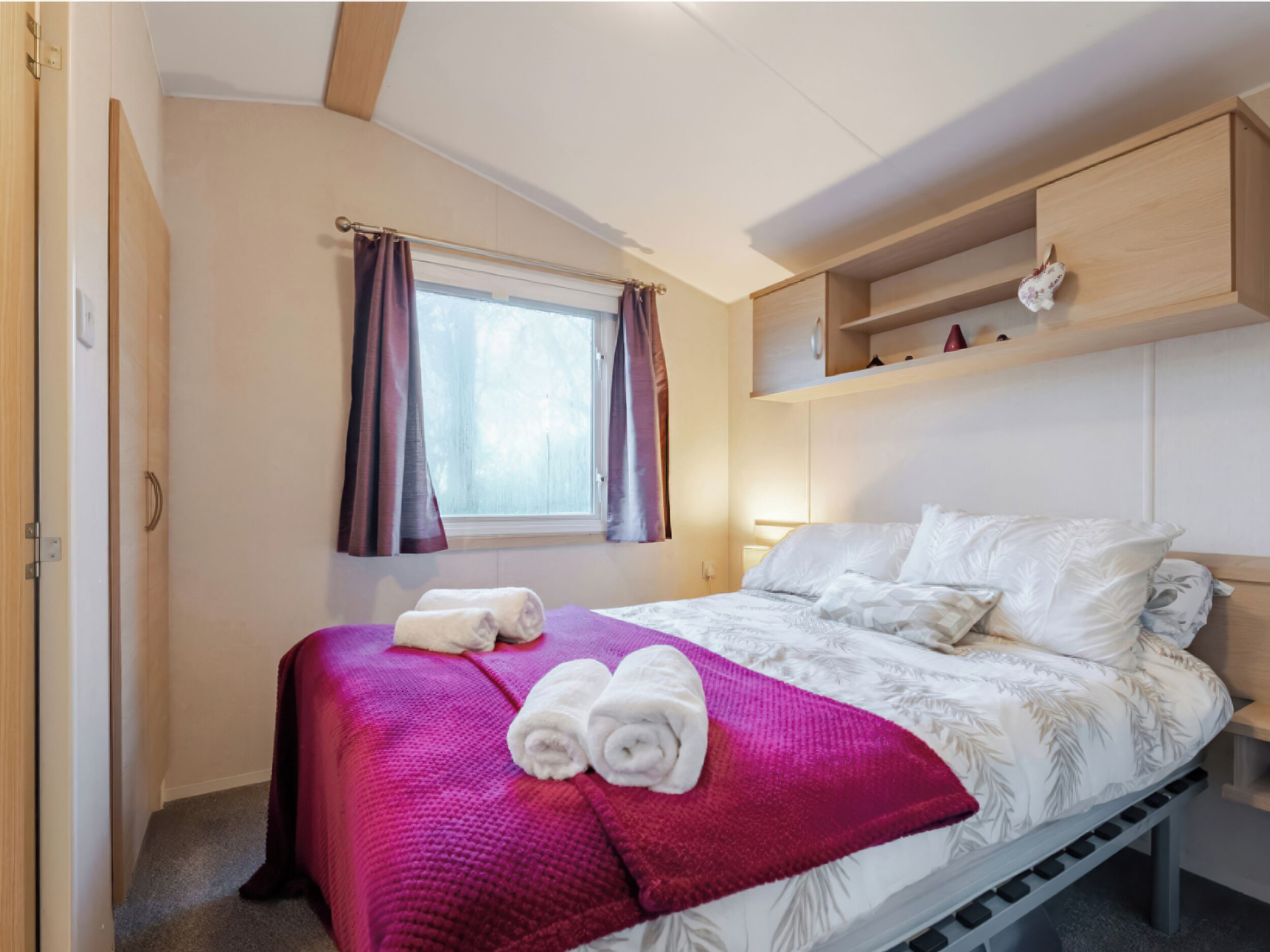 A bedroom with a bed, a window with curtains, a shelf with a decorative item, and a towel on the bed.