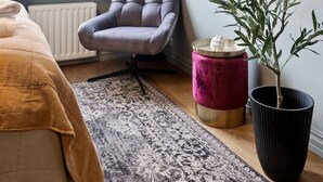 Een kamer met een raam, een grijze fauteuil, een kamerplant, een kleine tafel en een kamerbreed tapijt.