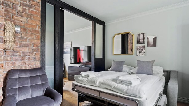 Een moderne slaapkamer met een bakstenen muur, een groot bed met witte lakens, een grijze fauteuil en een flatscreen televisie.