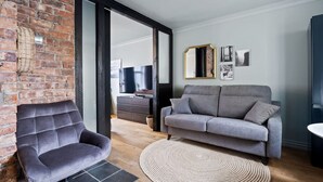 Een woonkamer met een bakstenen muur, een grijze bank, een grijze fauteuil, een geweven vloerkleed en een televisie.