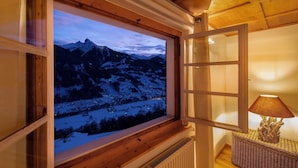 Ein Zimmer mit großem Fenster mit Blick auf eine verschneite Berglandschaft, einer Holzbank und einer Tischlampe.