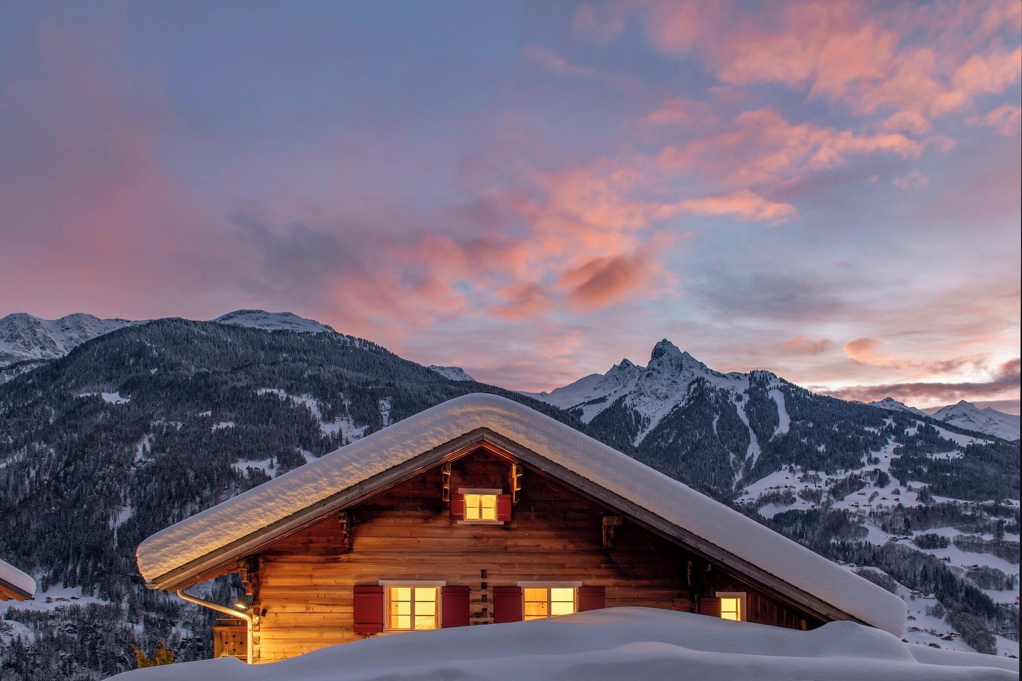 A wooden cabin with a sloped roof, surrounded by snow-covered mountains and a twilight sky.