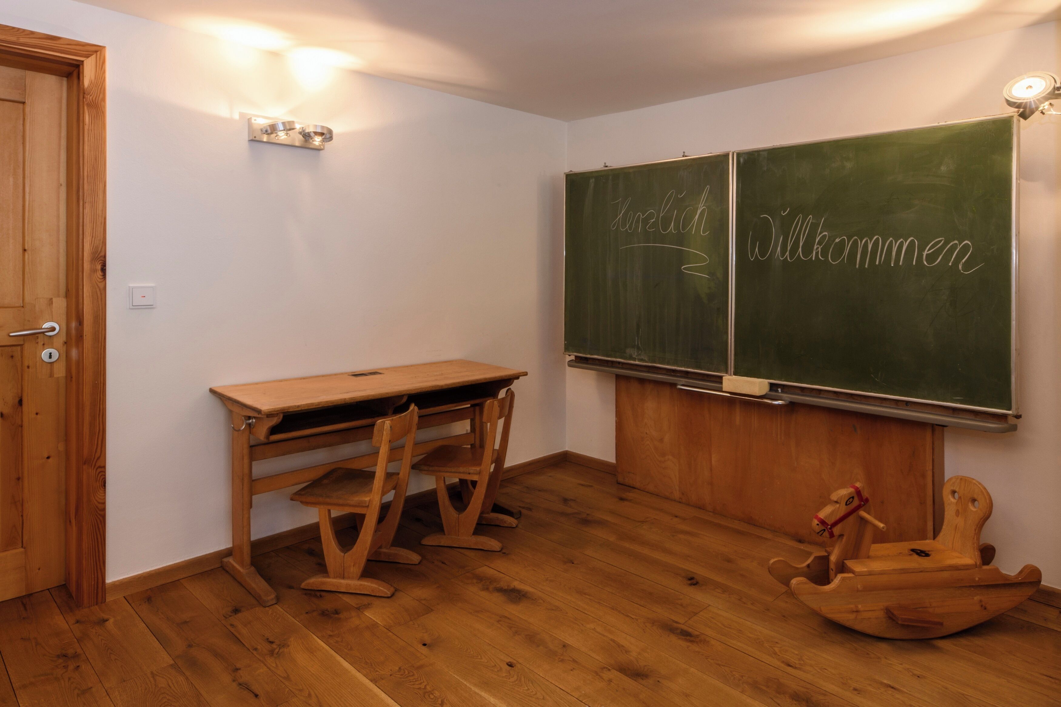 A classroom with a wooden desk, chairs, a chalkboard with 'Willkommen' written on it, and a wooden rocking horse.