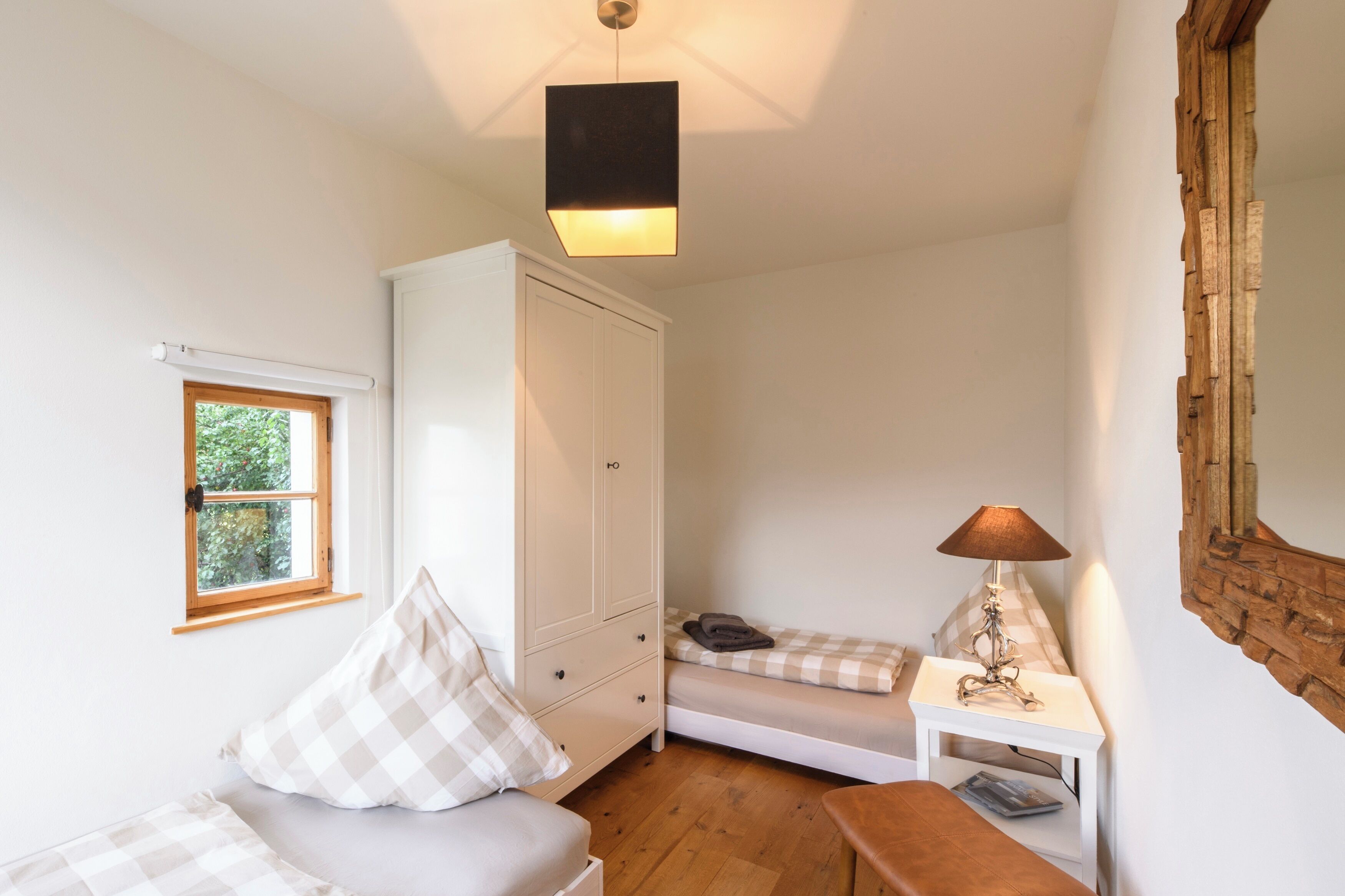 A small, well-lit room with a bed, a bedside table with a lamp, a wardrobe, and a window.
