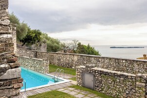 Una piscina de piedra con vistas al mar, rodeada de muros de piedra y un área con césped.
