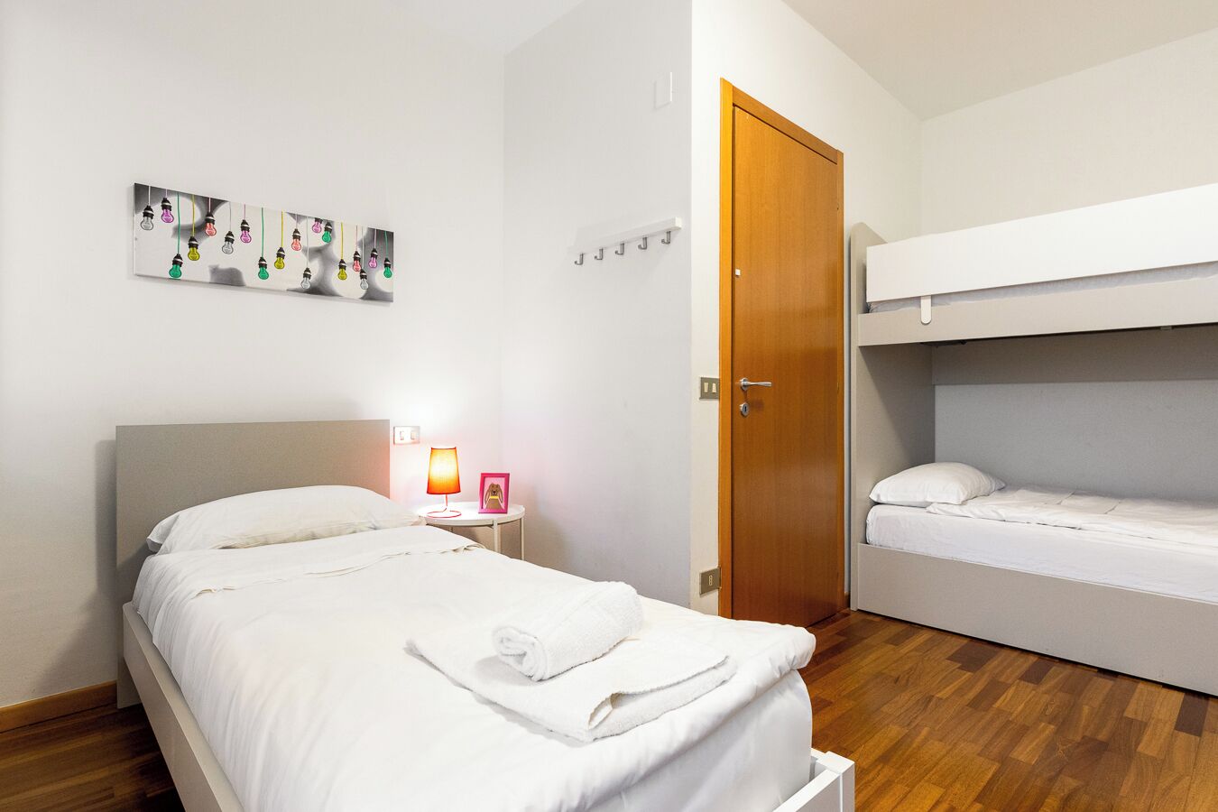 A hotel room with a single bed, a bedside table with a lamp and a picture frame, a wooden door, and a bunk bed.