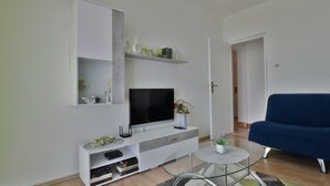 Ett modernt vardagsrum med en platt-TV, en blå soffa, ett soffbord i glas och en vit TV-möbel med hyllor.