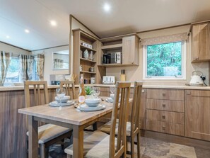 A modern kitchen with wooden cabinets, a dining table set for four, and a window with a view of greenery. - The But n Ben - Kelling Heath Holiday Park (Holt)