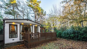 A mobile home with a deck, surrounded by trees and a leaf-covered ground. - The But n Ben - Kelling Heath Holiday Park (Holt)