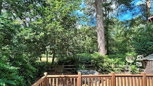 A wooden deck with a fence, surrounded by dense green foliage and a large tree. - The But n Ben - Kelling Heath Holiday Park (Holt)