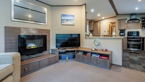 A modern living room with a flat-screen TV, a fireplace, and a kitchen area with an oven and microwave. - The But n Ben - Kelling Heath Holiday Park (Holt)