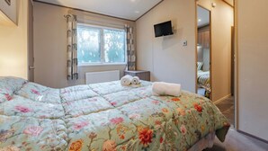 A bedroom with a bed, a window with curtains, a radiator, a TV mounted on the wall, and a door leading to another room. - The But n Ben - Kelling Heath Holiday Park (Holt)