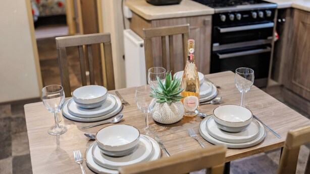 A dining table set with plates, glasses, cutlery, a bottle, and a decorative plant. - The But n Ben - Kelling Heath Holiday Park (Holt)