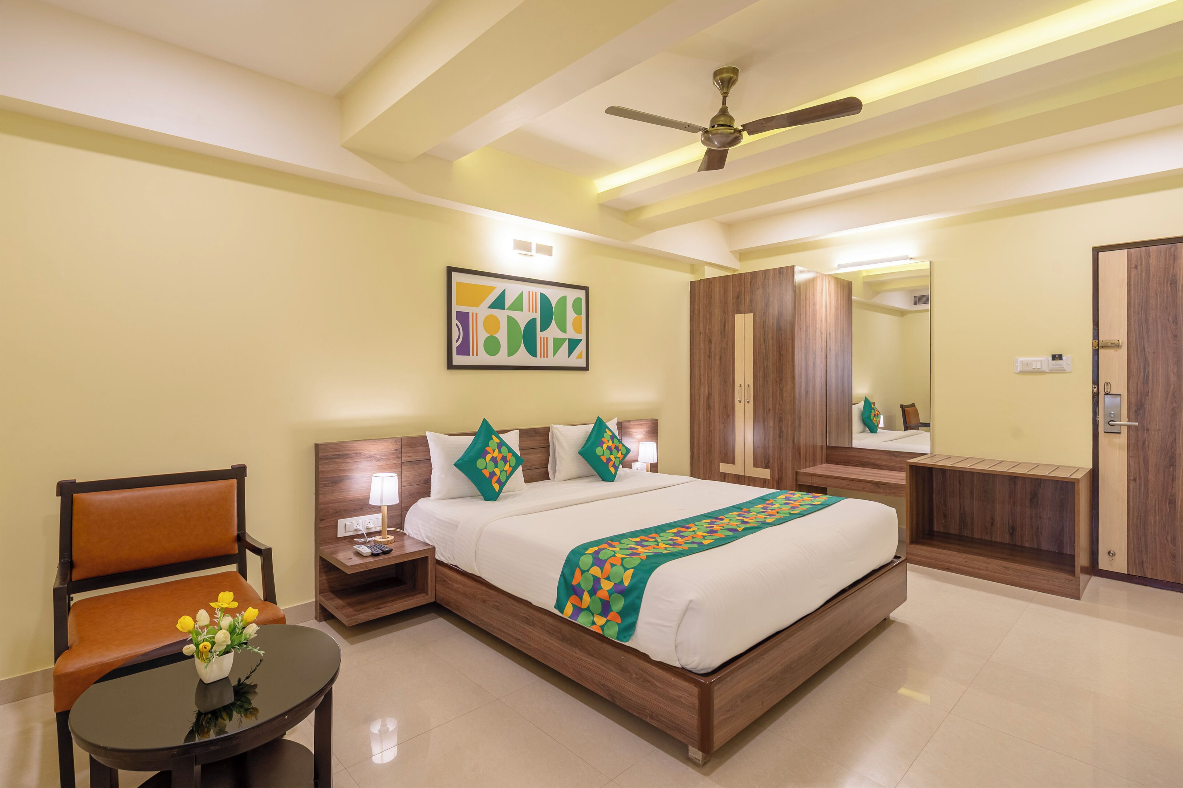 A hotel room with a bed, a chair, a small table with flowers, a nightstand, a ceiling fan, and a mirror.