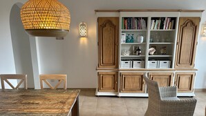 Zona de menjador amb taula i cadires de fusta, una cadira de vímet i una biblioteca amb llibres i objectes decoratius.