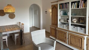 Zona de menjador amb taula i cadires de fusta, una cadira de vimetla, una biblioteca amb objectes decoratius i una làmpada penjant.